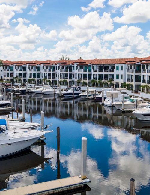 A waterfront marina with yachts moored along calm canals beside a row of white, red-roofed resort buildings under sunny clouds.