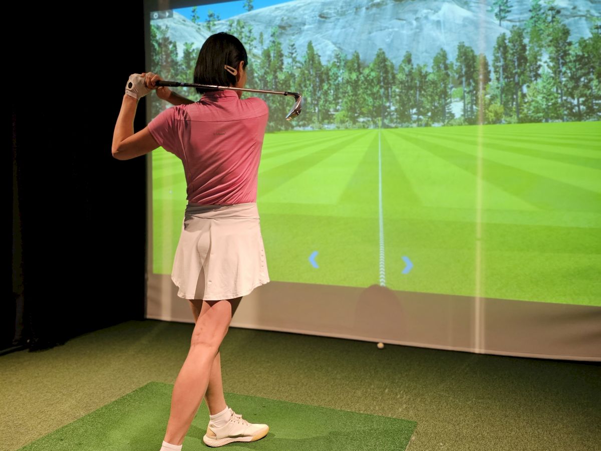 A woman swings a golf club in an indoor simulator, aiming at a projected course on the big screen.