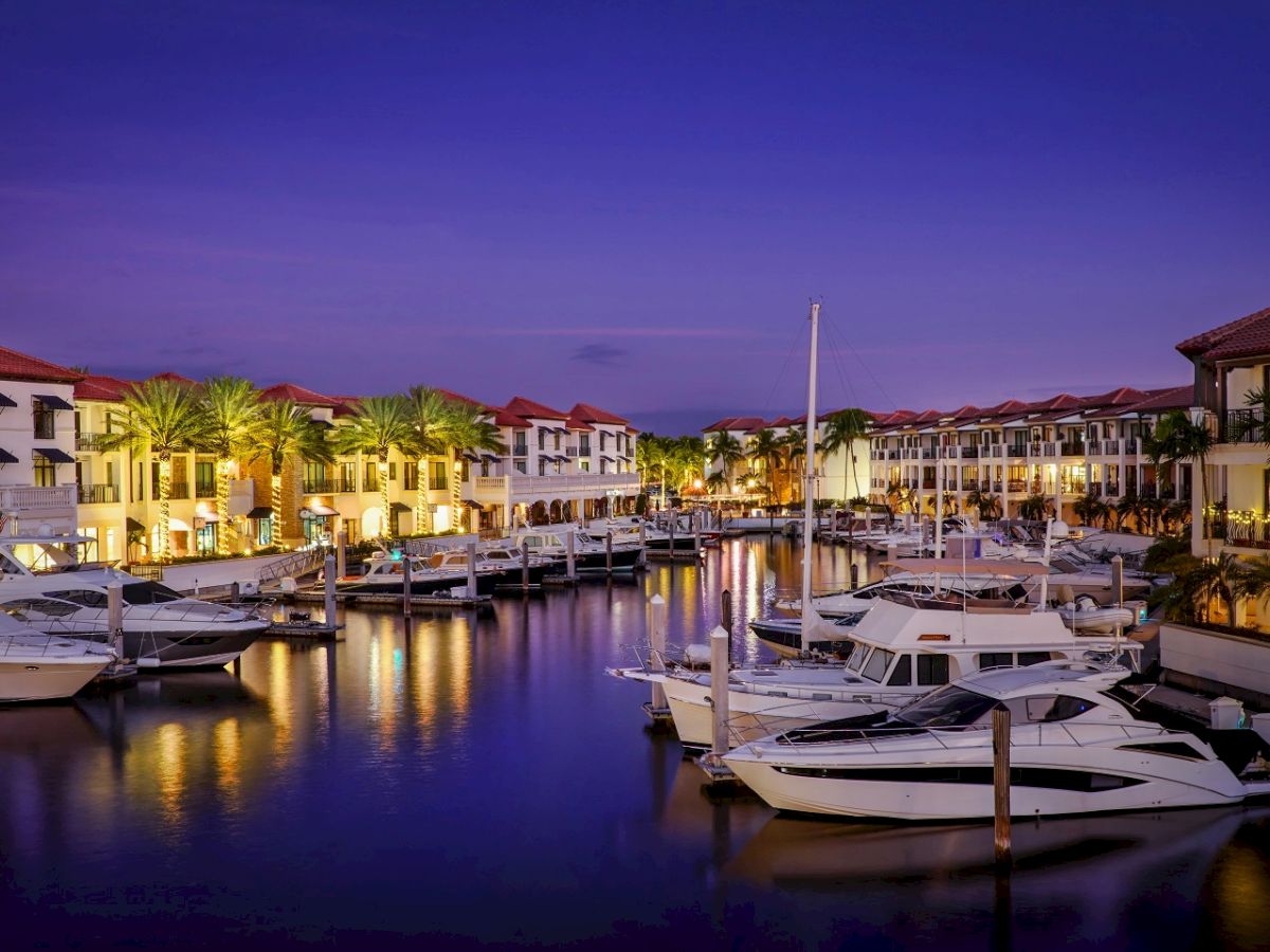 Dusk marina scene with yachts docked, palm-lined red-roof resort buildings lit up, calm purple sky reflecting on the water.