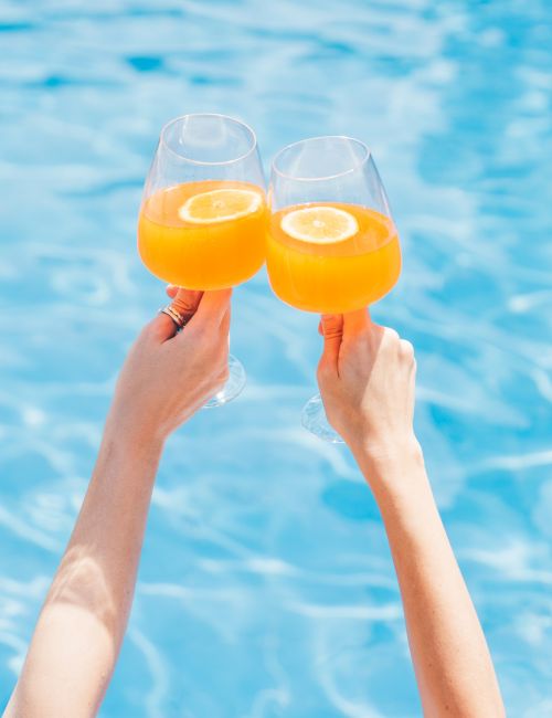 Two hands clink glasses of orange juice with lemon slices, enjoying a refreshing drink poolside in the summer sun.