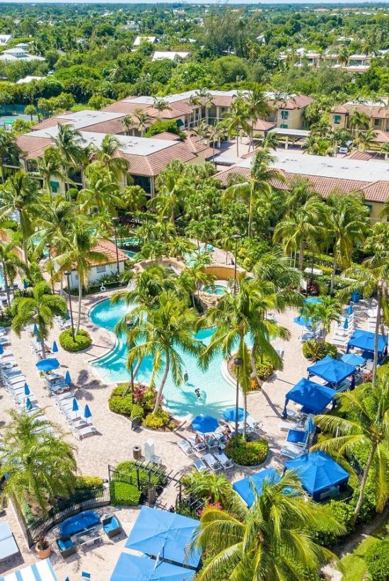 Aerial view of a tropical resort with multiple swimming pools, blue umbrellas, and lush palm trees surrounding the villas.