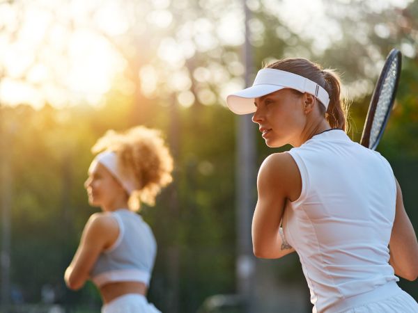 A woman in a white visor swings a tennis racket beside a teammate on an outdoor court at golden sunset.
