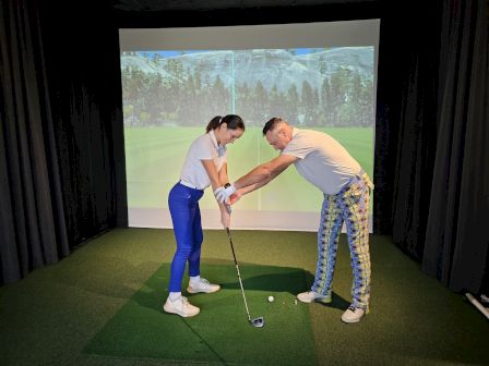 A golfer is being coached in an indoor simulator, with a club and ball on green turf before a big screen.