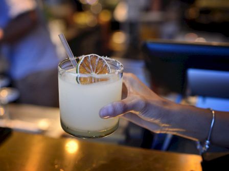 A bartender&rsquo;s hand holds a frosty cocktail in a rocks glass with ice, a dried orange wheel, and a straw.