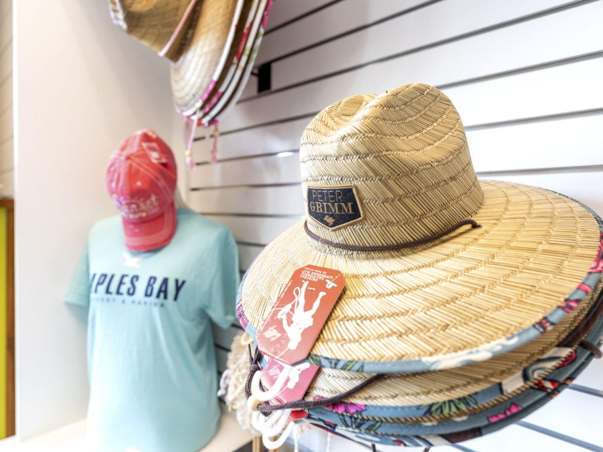 A store display shows straw hats stacked on a shelf next to a blue &ldquo;Isles Bay&rdquo; T-shirt mannequin; pink items hang nearby.