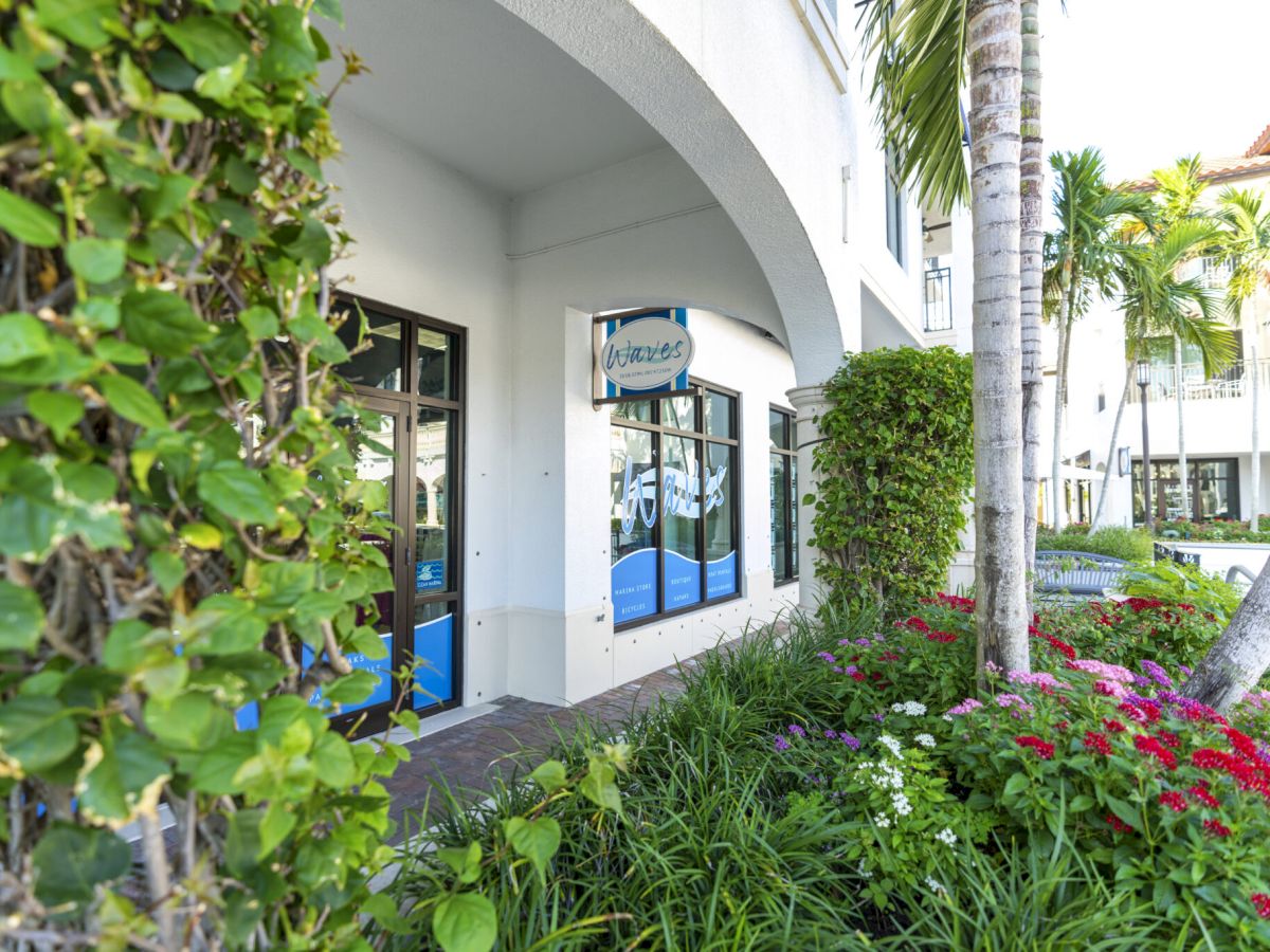 A sunny storefront with arched entrance, lush landscaping, palm trees, and flowering bushes along a white building.