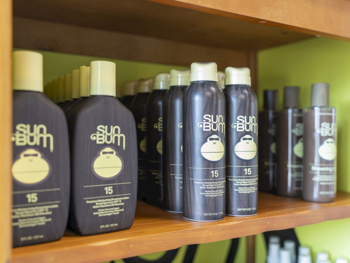 Arranged bottles of Sun Bum sunscreen on a shelf, featuring dark labels and a sun logo, with SPF 15 and varied sizes, neatly lined up.