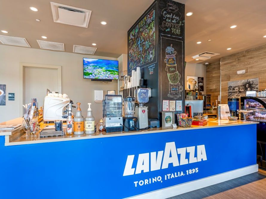 A bright Lavazza caf&eacute; counter with a blue front, coffee machines, syrups, and a viewing TV in a modern lounge.