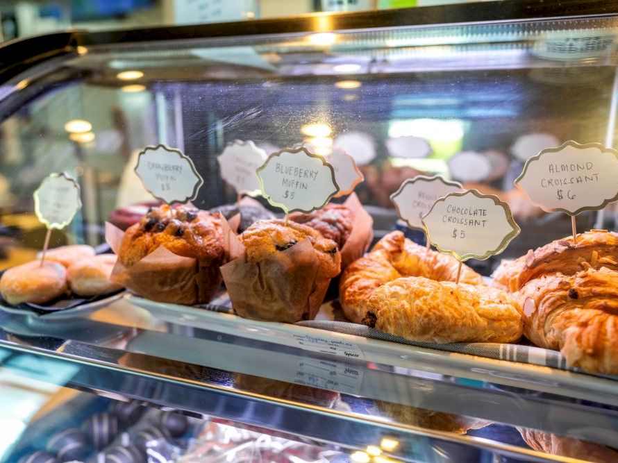 A glass display case of assorted pastries&mdash;muffins, croissants, and Danish pastries, labeled with small signs, in a cafe setting.